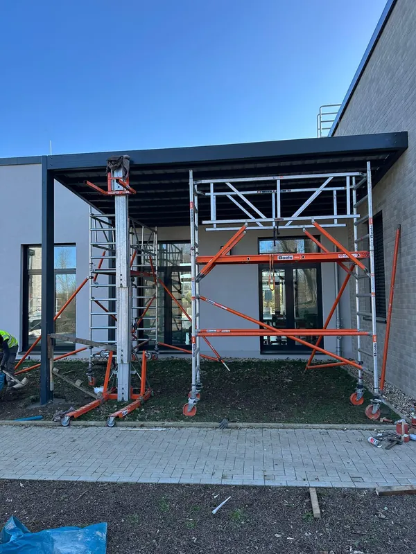 Dunkle Metallstützen und ein Dach vor einer Hausfassade; darunter stehen Montagegerüste.